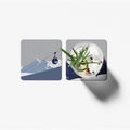 Two square coasters with mountain and ski lift design on one, and cocktail with garnish on the other, on a white background.
